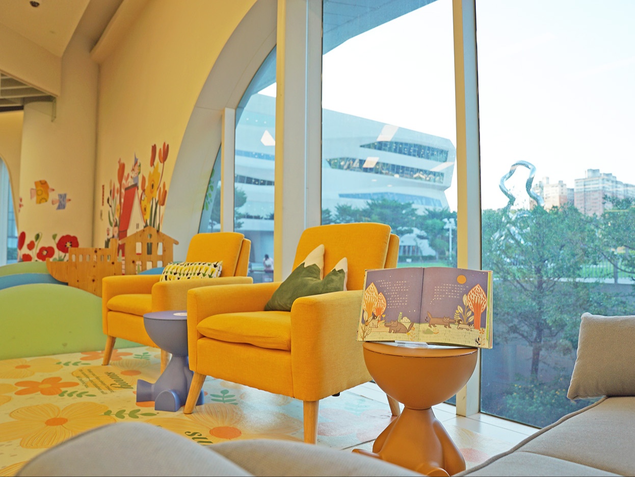 一樓「親輕讀」閱讀角，落地窗的藍天、綠樹、光影交疊，營造明亮舒適的空間氛圍。父母陪孩子共讀一本書，在自然中享受溫暖的親子時光。閱讀成為親子最溫暖的日常風景